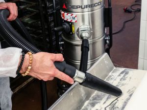 Close-up of a Depureco vacuum nozzle collecting fine powder from a metal tray; stainless industrial vacuum in background.