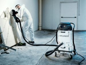 Worker sanding drywall with a hose-connected Depureco dust extractor capturing fine silica dust.
