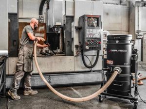 Technician vacuuming metal chips and coolant from a CNC machine with a wheeled sump vacuum and hose.