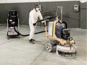 Worker in PPE operating a concrete grinder connected by hose to a mobile Depureco dust extractor for floor prep.